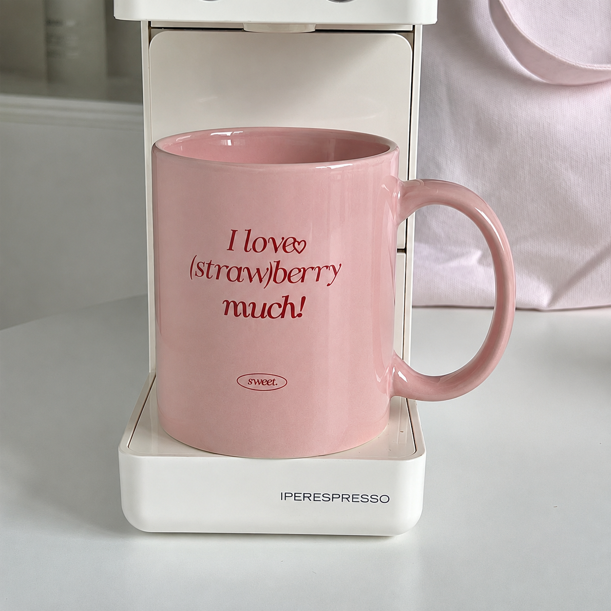 A pink ceramic coffee mug with the text 'I love (strawberry) much!' in red script, shown sitting inside a white IperESPRESSO coffee machine. The background is a soft, out-of-focus light pink fabric.