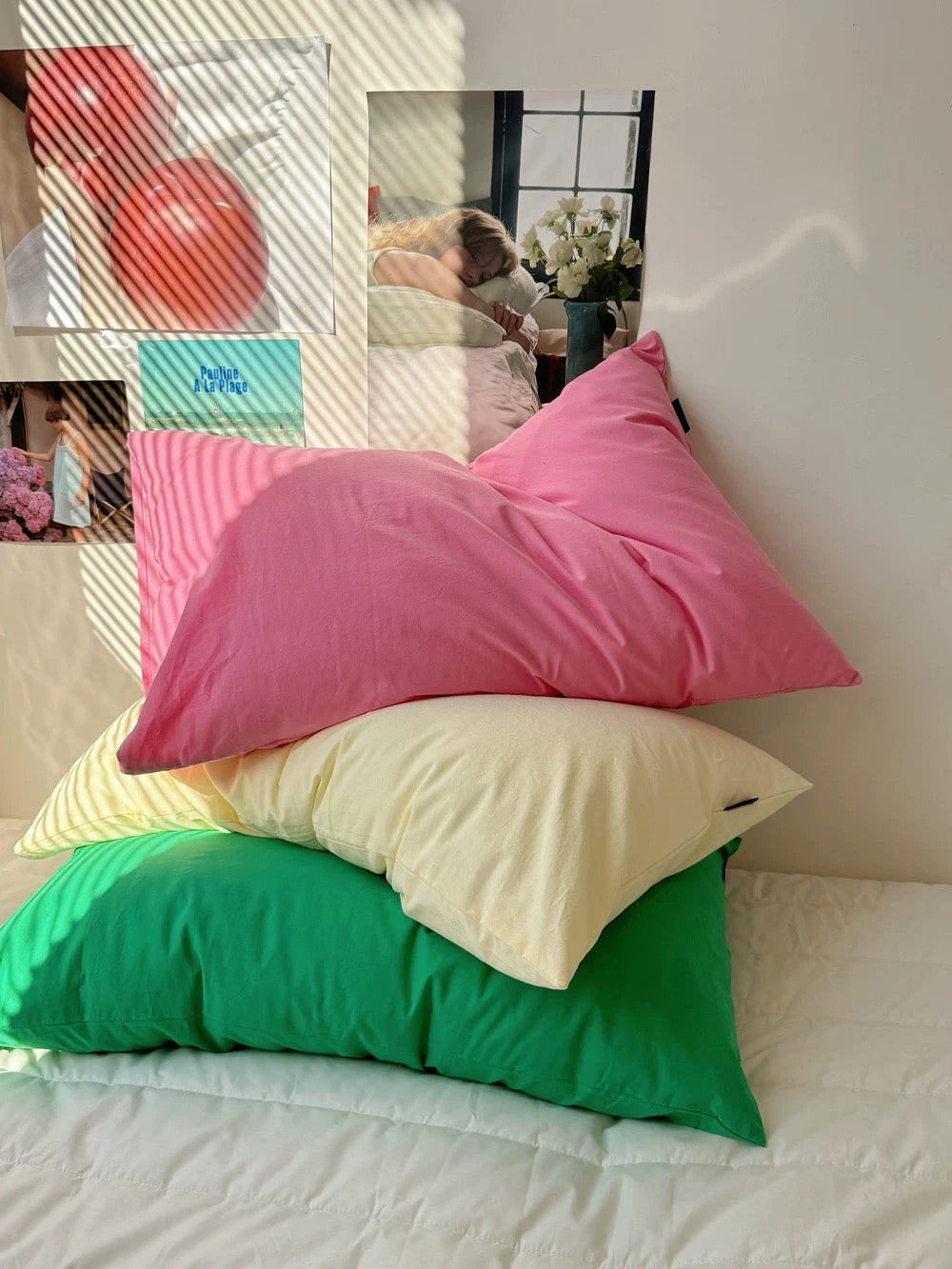 Three colorful pillows stacked on a bed: a bright pink pillow on top, a pale yellow pillow in the middle, and a green pillow at the bottom, set against a white wall with decorative posters and window light.