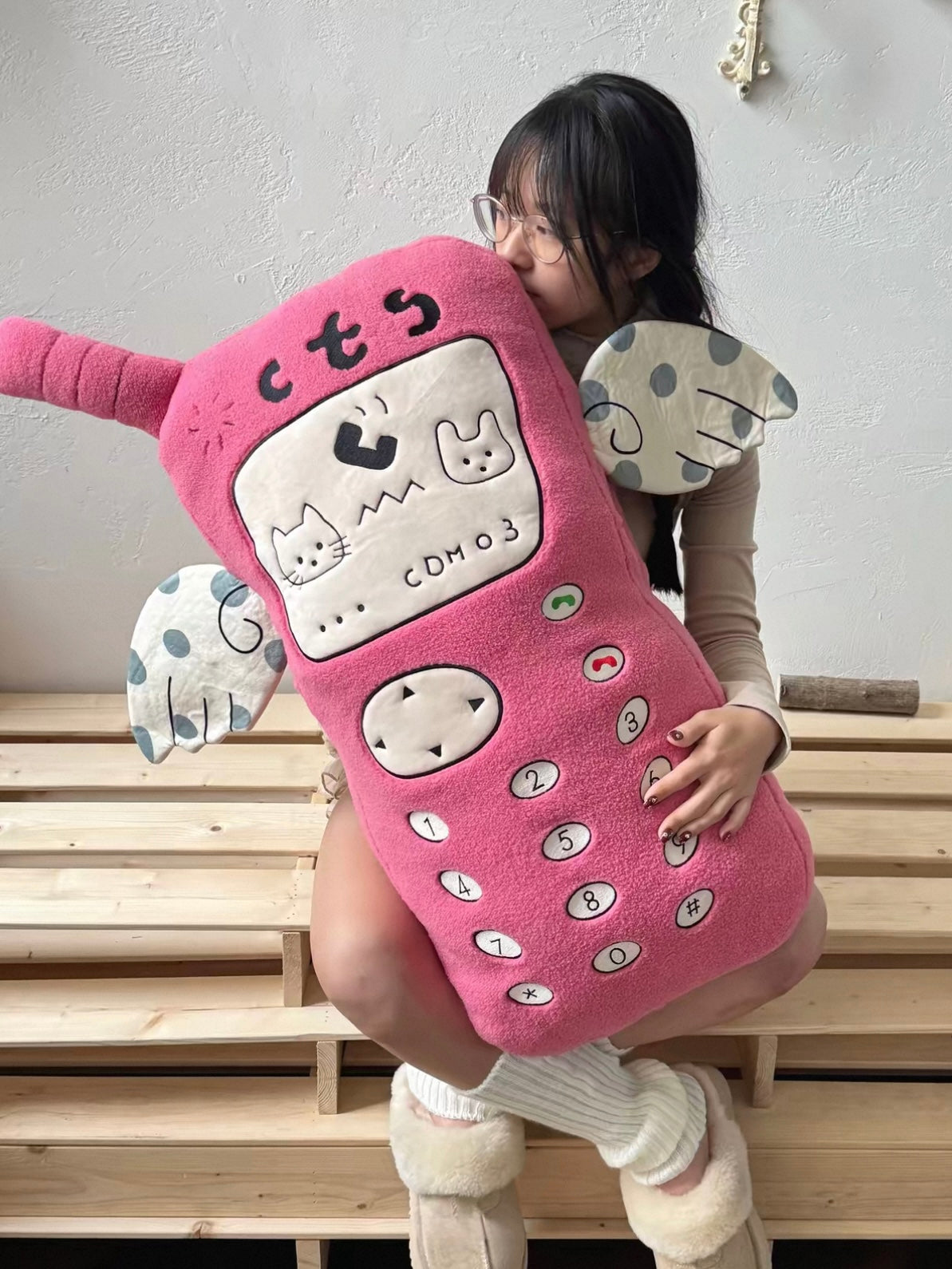 A person holds a pink phone plush close to their body, with the embroidered keypad and spotted arms visible.