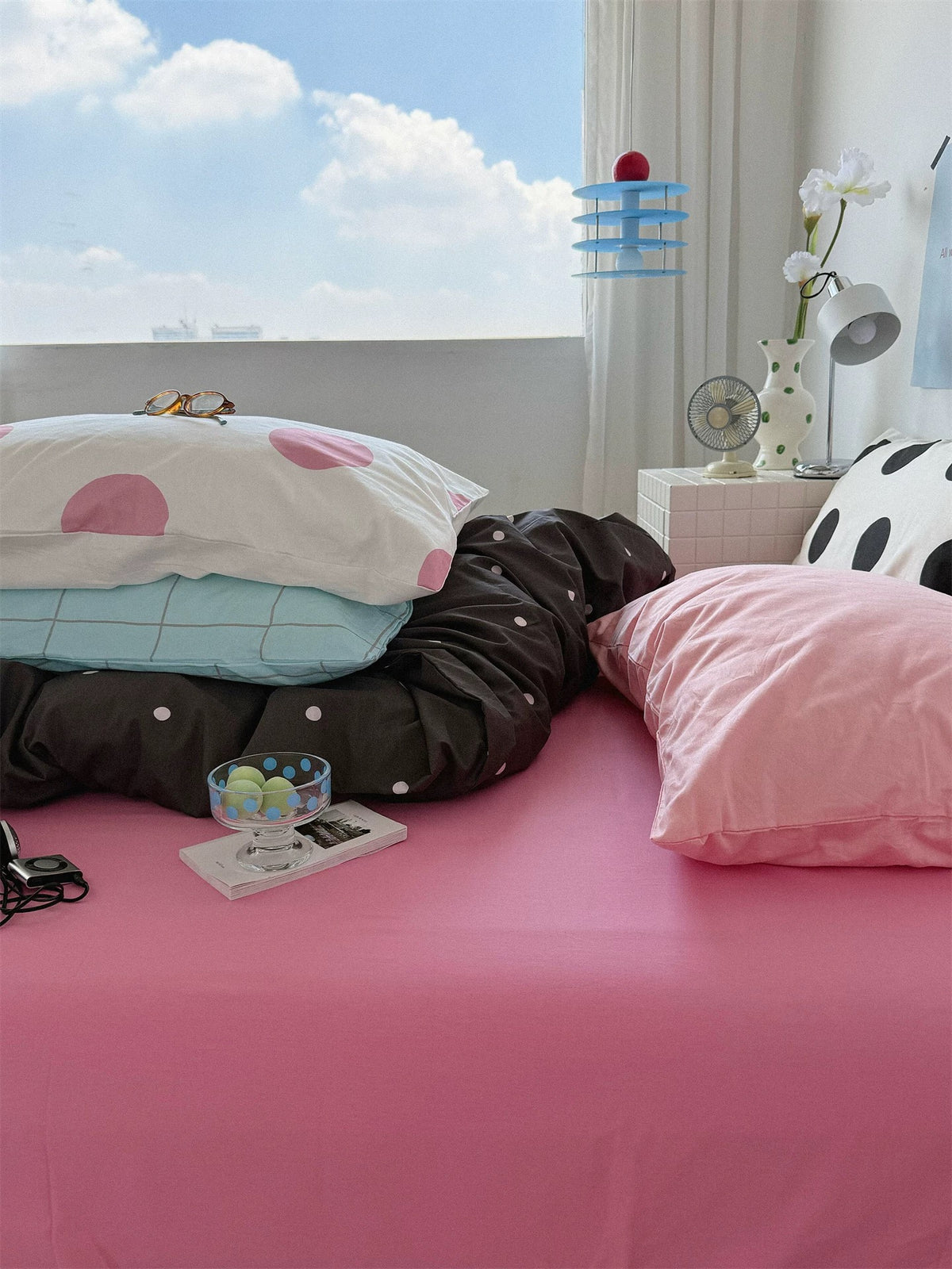 A pink sheet stretches across the bed beside a window, with dotted bedding and stacked pillows near the head.