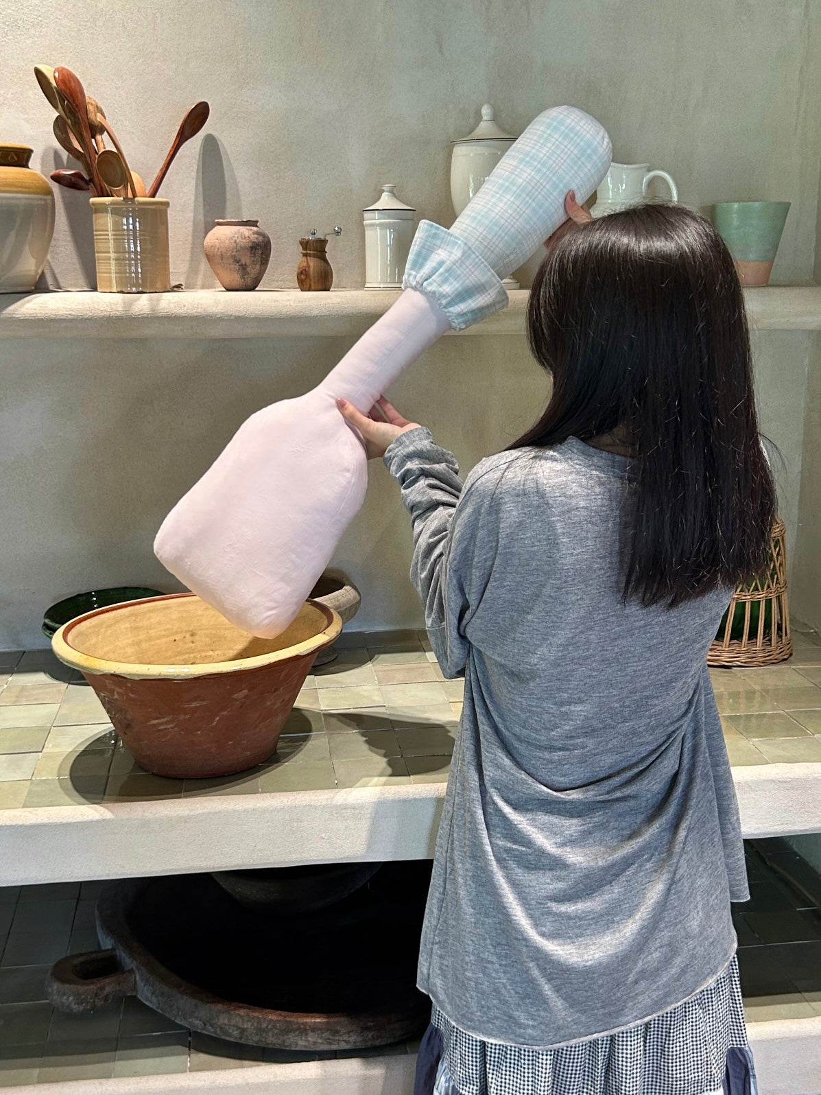 A person holds a pink spatula pillow across one shoulder, showing the pale blue checked handle and ruffled cuff.