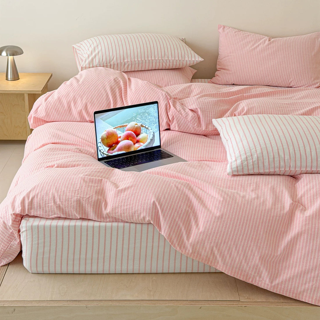 A bed dressed in a light pink and white striped bedding set, including a duvet cover, two pink pillows, and two striped pillows, set in a minimalist room with a wooden platform bed and a small side table.