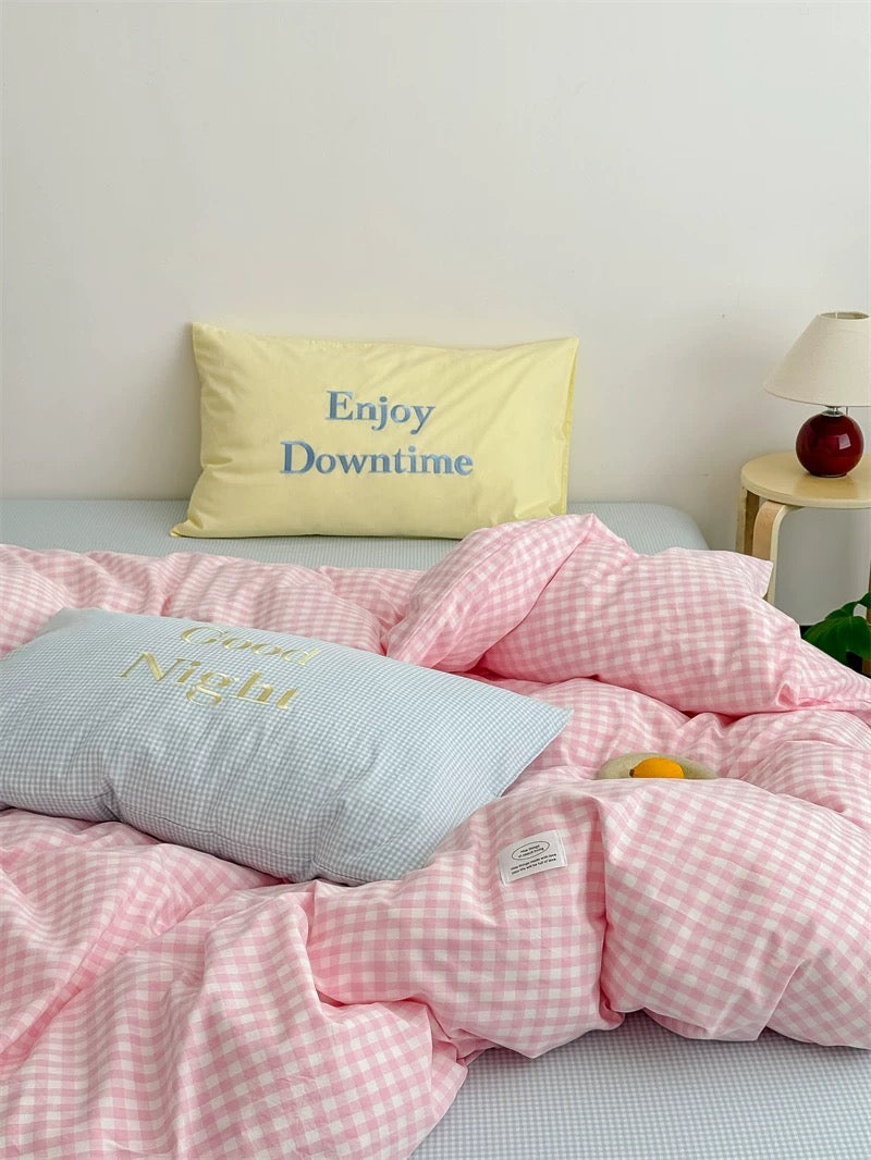 Bed with pink checkered duvet and pillows with text, nightstand with lamp in the background