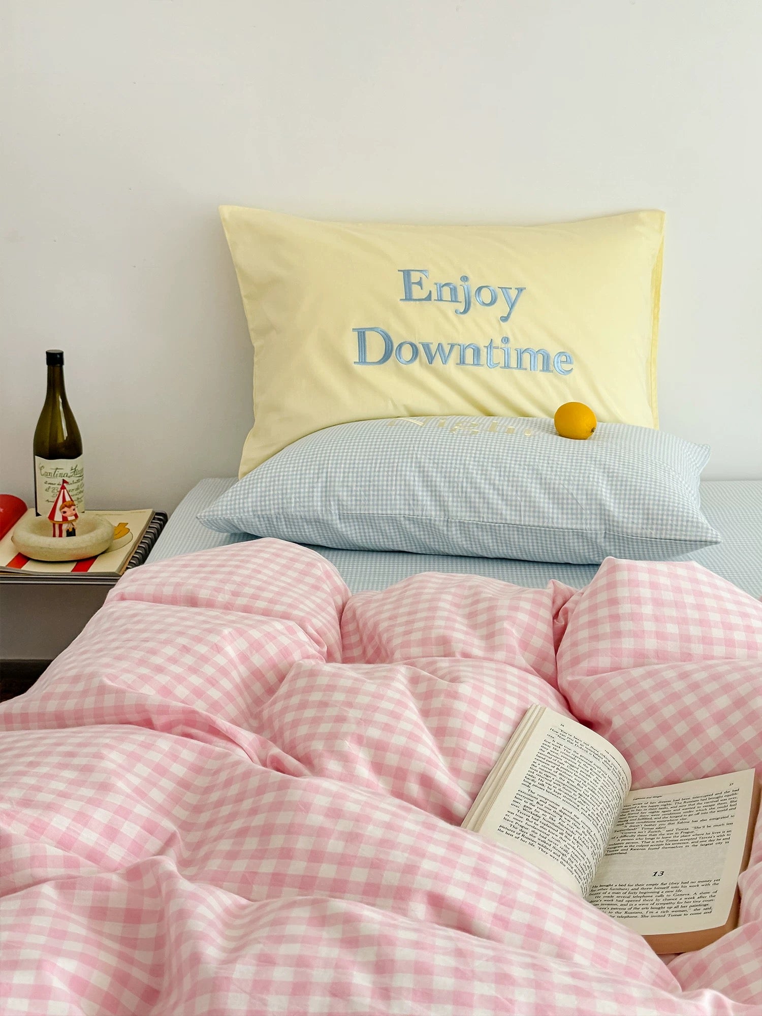 Bed with pink checkered comforter, yellow pillow with 'Enjoy Downtime' text, and a bottle on a nightstand.