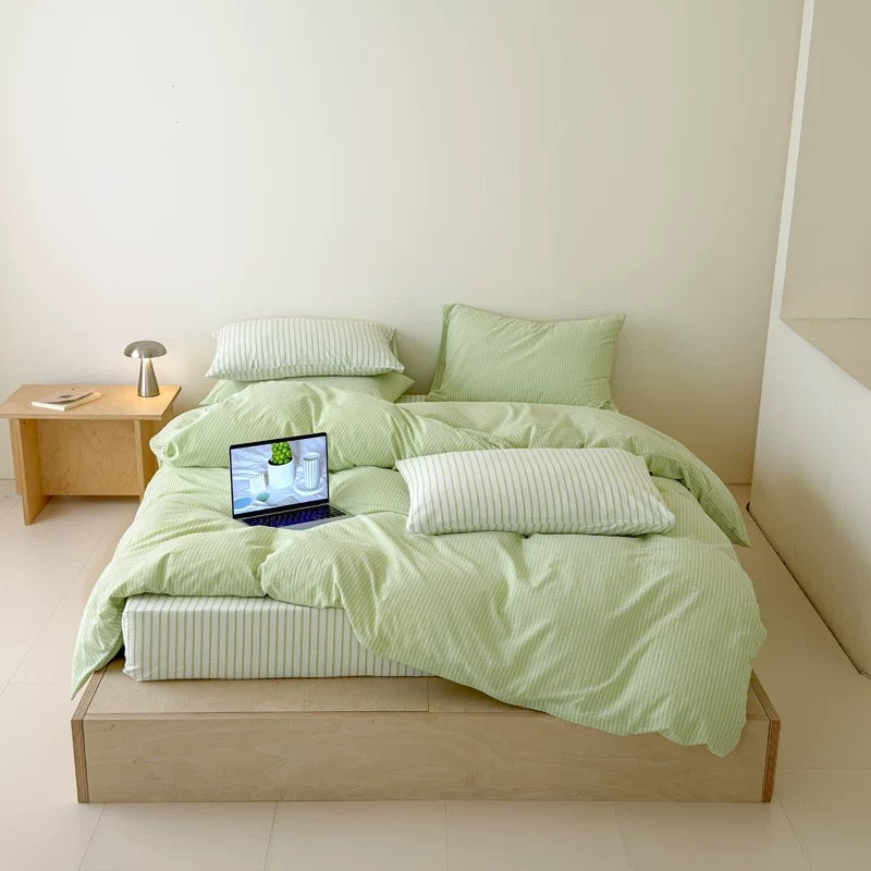 A bed dressed in light green bedding with matching pillows and a striped pillow, set in a minimalist room with a wooden bed frame, side table, and laptop on the mattress.