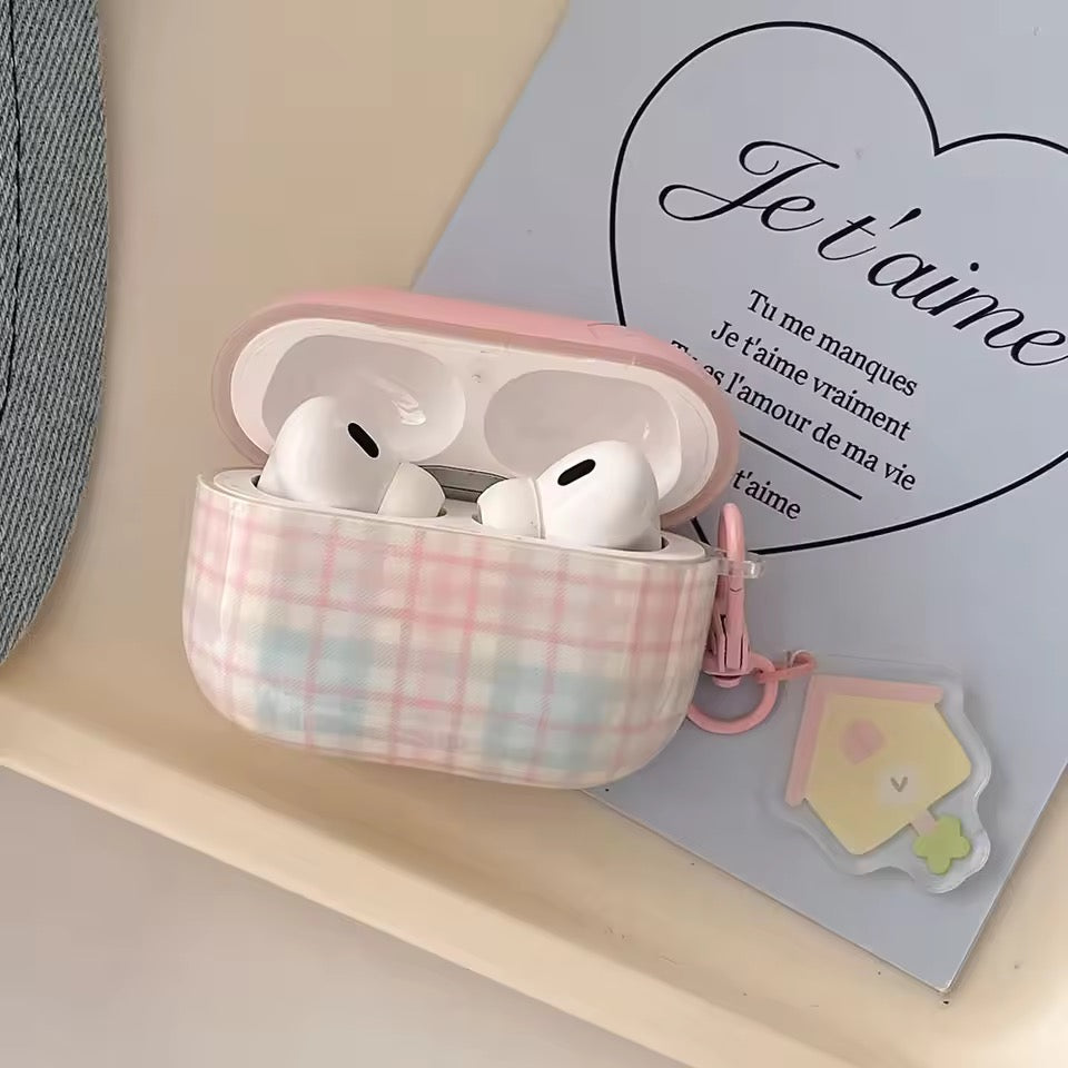 Wireless earbuds in a pink plaid case on a light surface with a heart-shaped 'Je t'aime' card in the background.