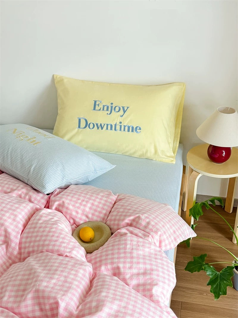 Bed with pink checkered duvet and yellow pillow with 'Enjoy Downtime' text, side table with lamp.