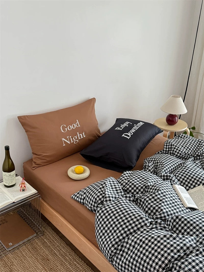 Bed with checkered duvet, pillows, and a small table with a bottle and fruit.