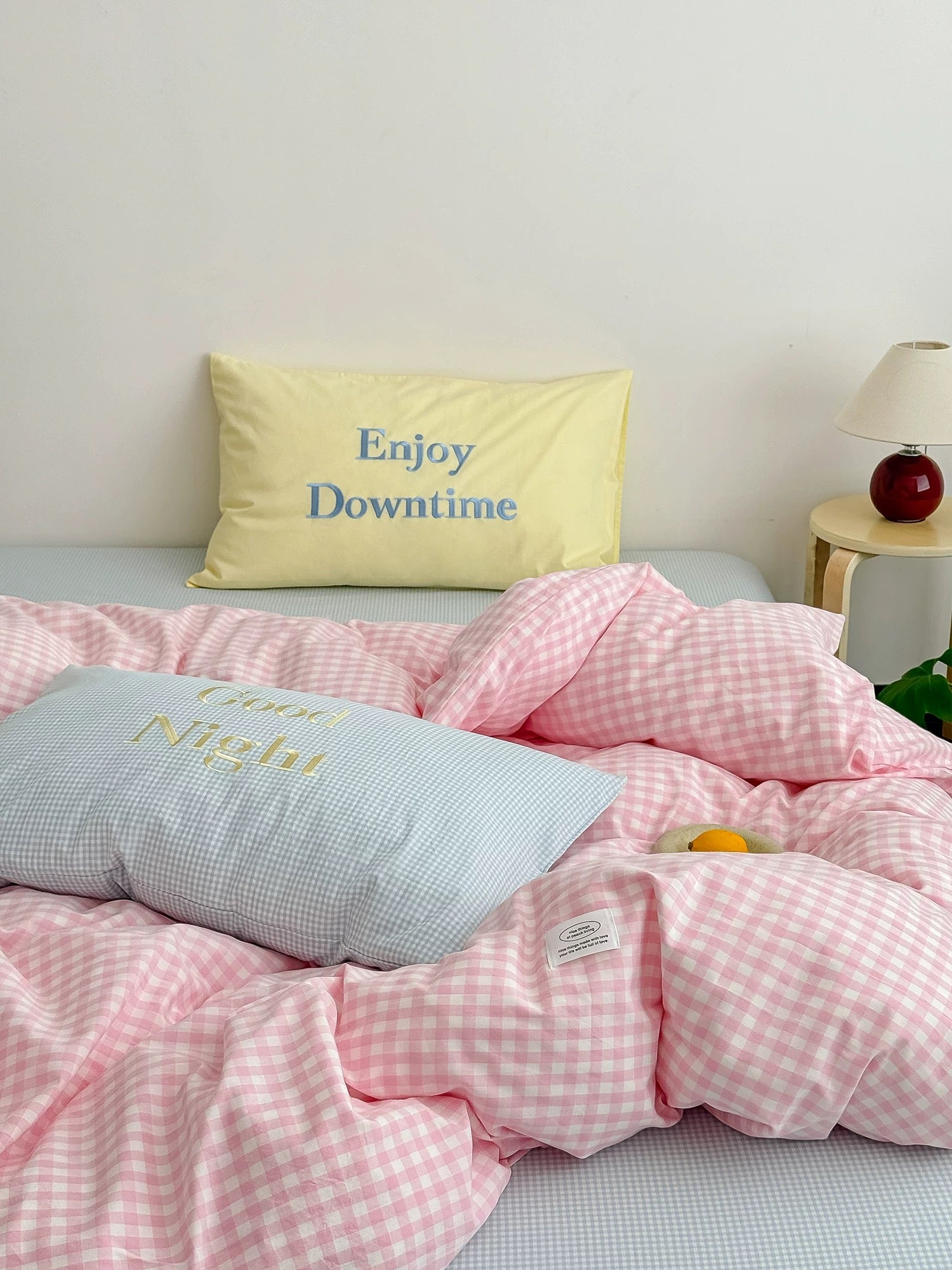 Bed with pink checkered duvet and pillows, one with 'Enjoy Downtime' text, in a bedroom setting.