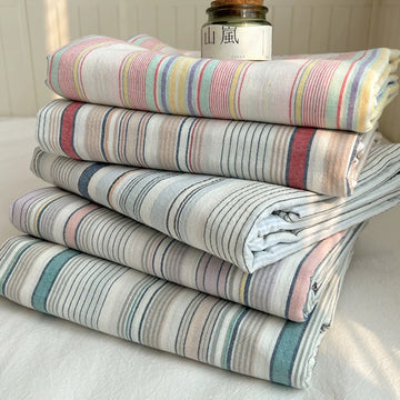 A stack of five folded striped textiles, likely towels, blankets, or sheets, in soft pastel colors including pink, blue, yellow, and green on a white base. A small glass jar with a white label sits on top.