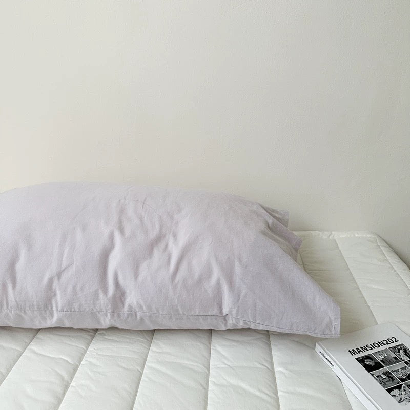A pale purple gray pillowcase rests against a white headboard with an open book at the foot of the bed.