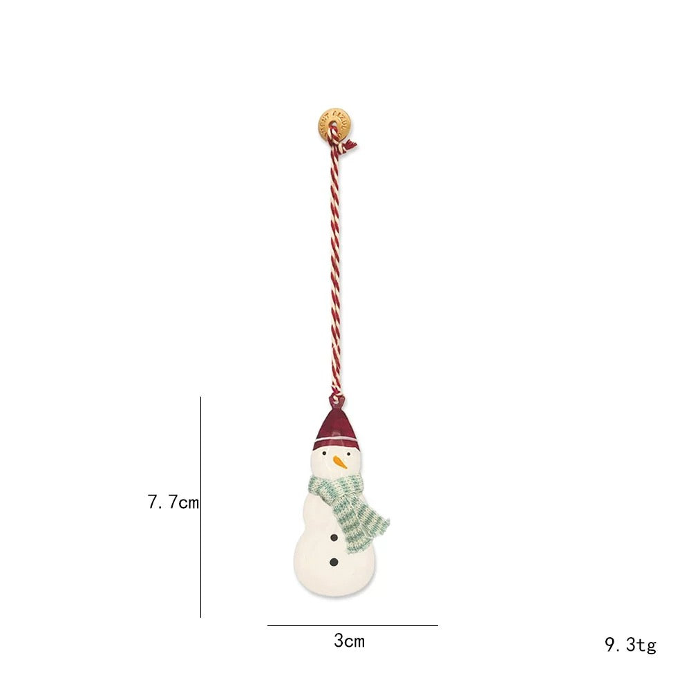 A snowman Christmas ornament wears a red pointed hat and green checked scarf above black buttons.