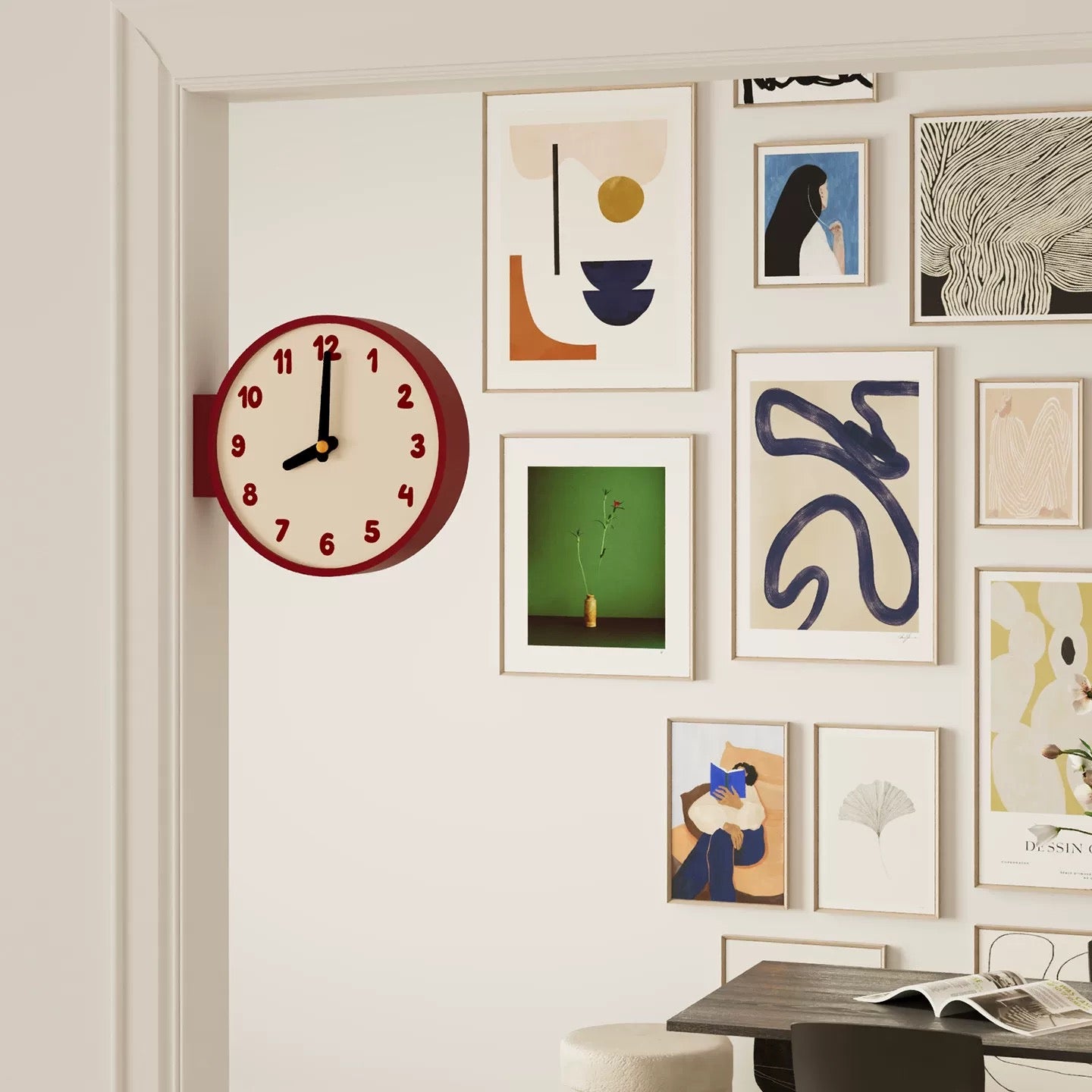 A round wall clock with a dark red frame and cream face hangs on a white wall beside a gallery wall of framed abstract and colorful art prints. The clock has black hands and red numbers.