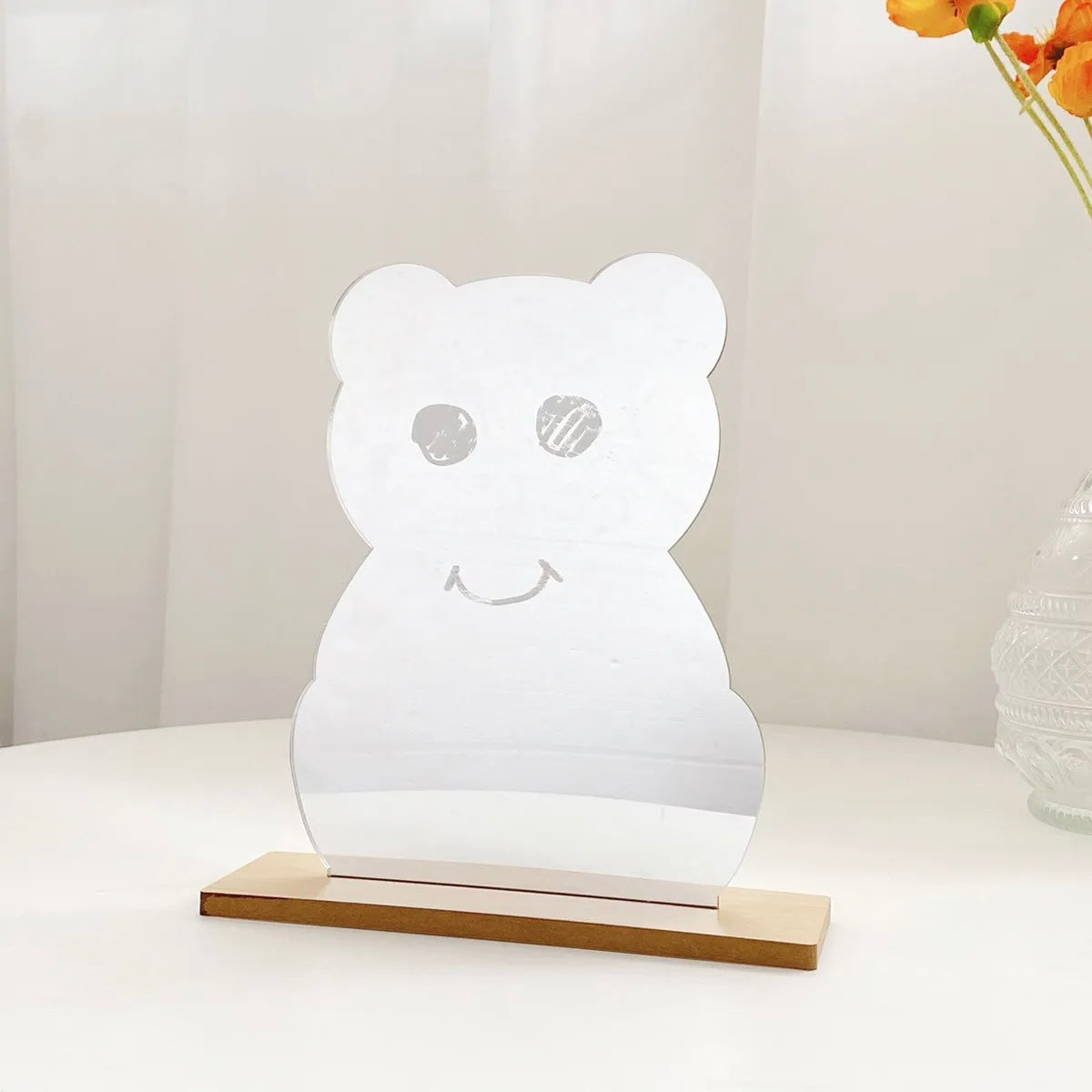 A clear acrylic bear-shaped mirror with a wooden base, displayed on a white table against a soft, light background.