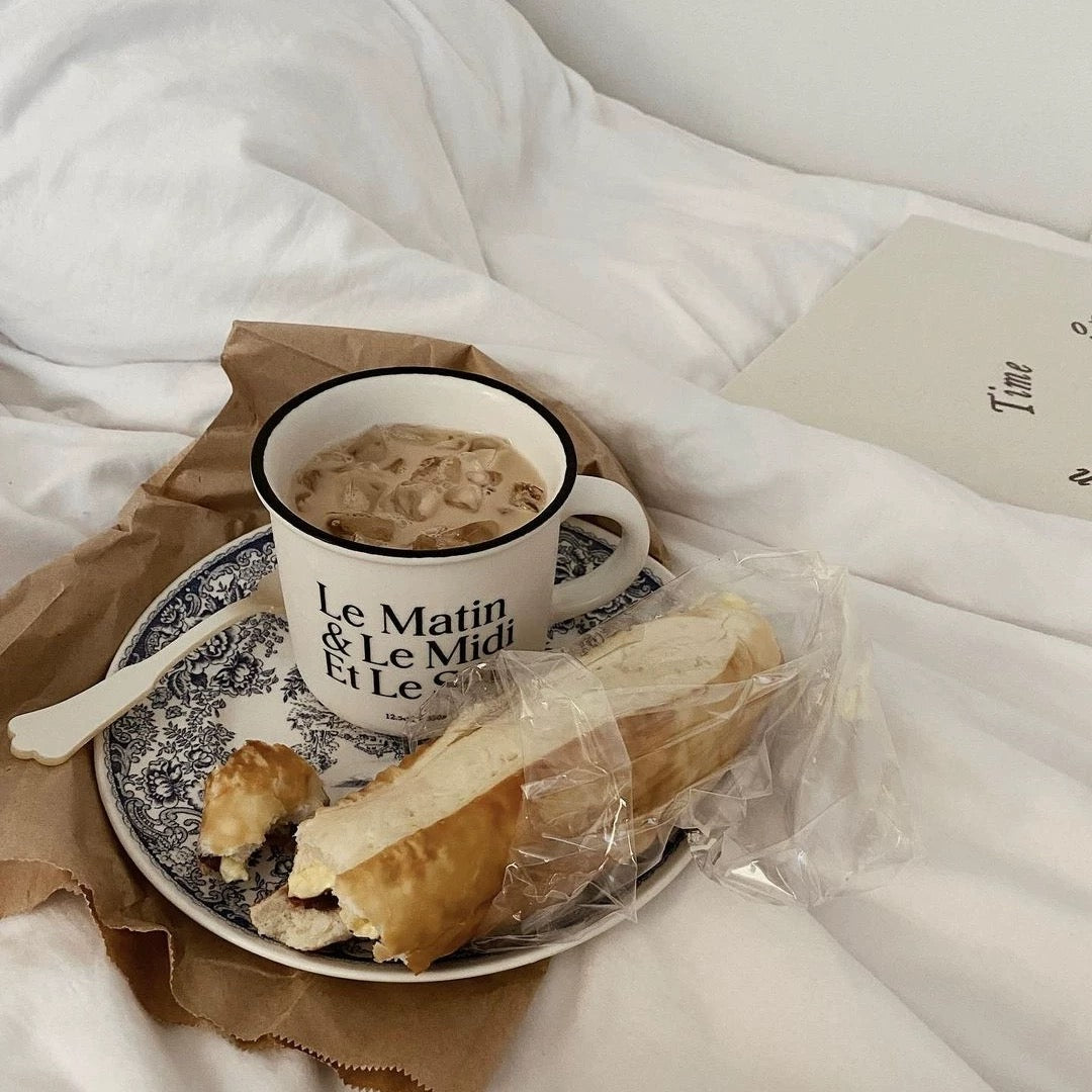 A white ceramic mug with black text reading 'Le Matin & Le Midi Et Le Soir' sits on a blue-and-white patterned saucer with a spoon, next to a baguette and pastry on a bed with white bedding.