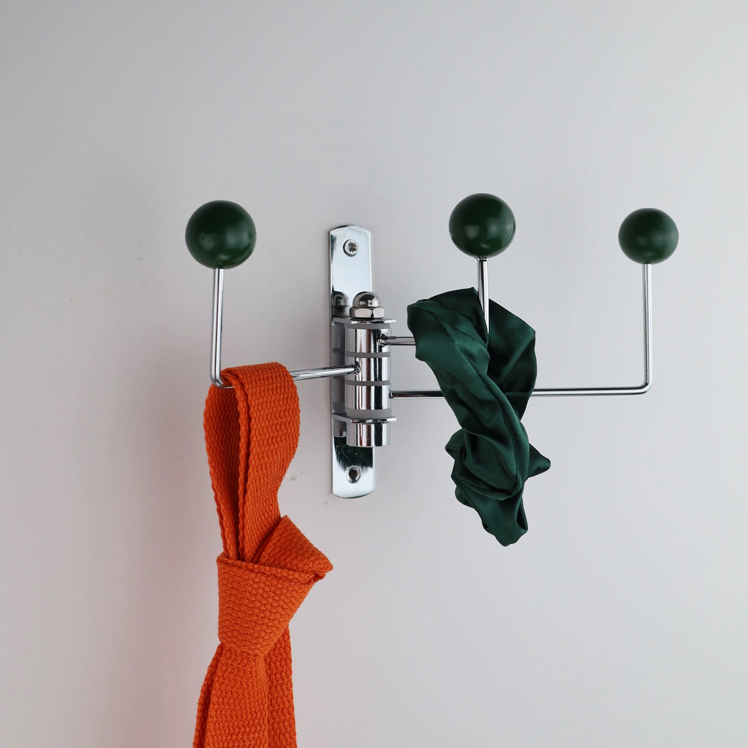 A wall-mounted three-hook coat rack with a polished silver metal frame and dark green spherical hook knobs. An orange woven scarf hangs from the left hook, a dark green fabric item hangs from the center-right horizontal bar, and a third dark green scarf is draped over the right vertical hook, all against a plain light gray wall.