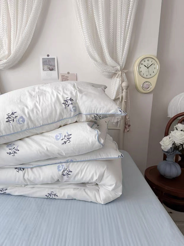 Stack of white bedding with floral embroidery on a bed in a bedroom setting.