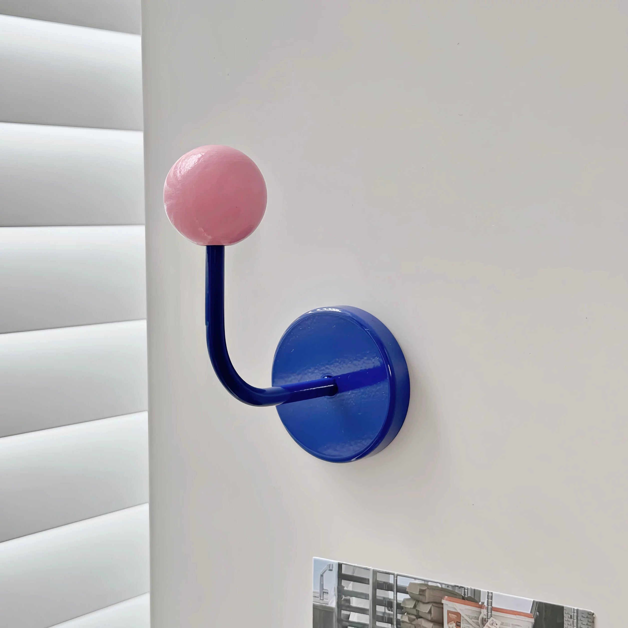A modern wall-mounted coat rack hook with a round pink top and a curved blue arm attached to a round blue wall plate, shown mounted on a white wall next to window blinds.