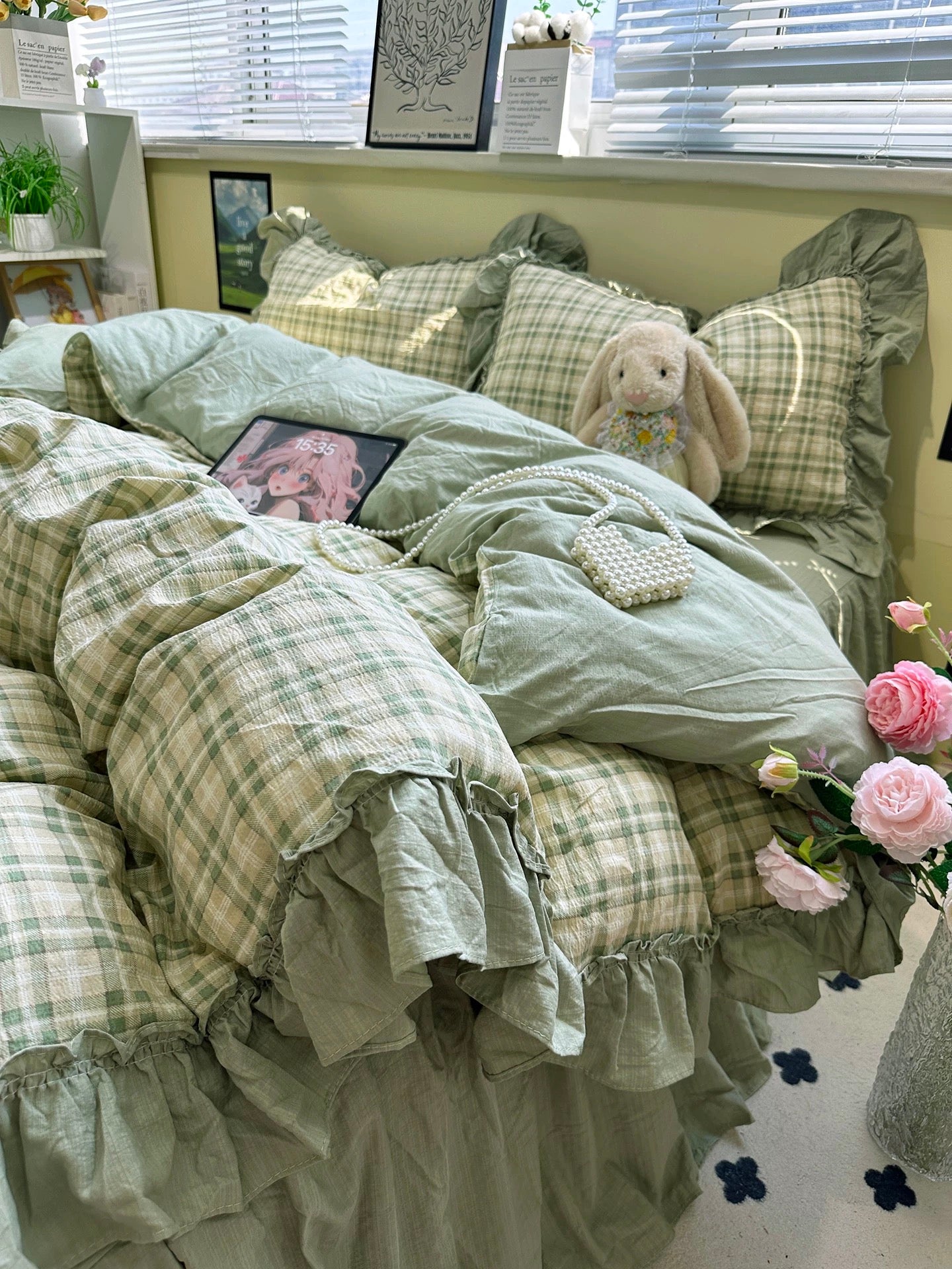 Bed with green plaid bedding, a teddy bear, and flowers in a room with a window and plants.