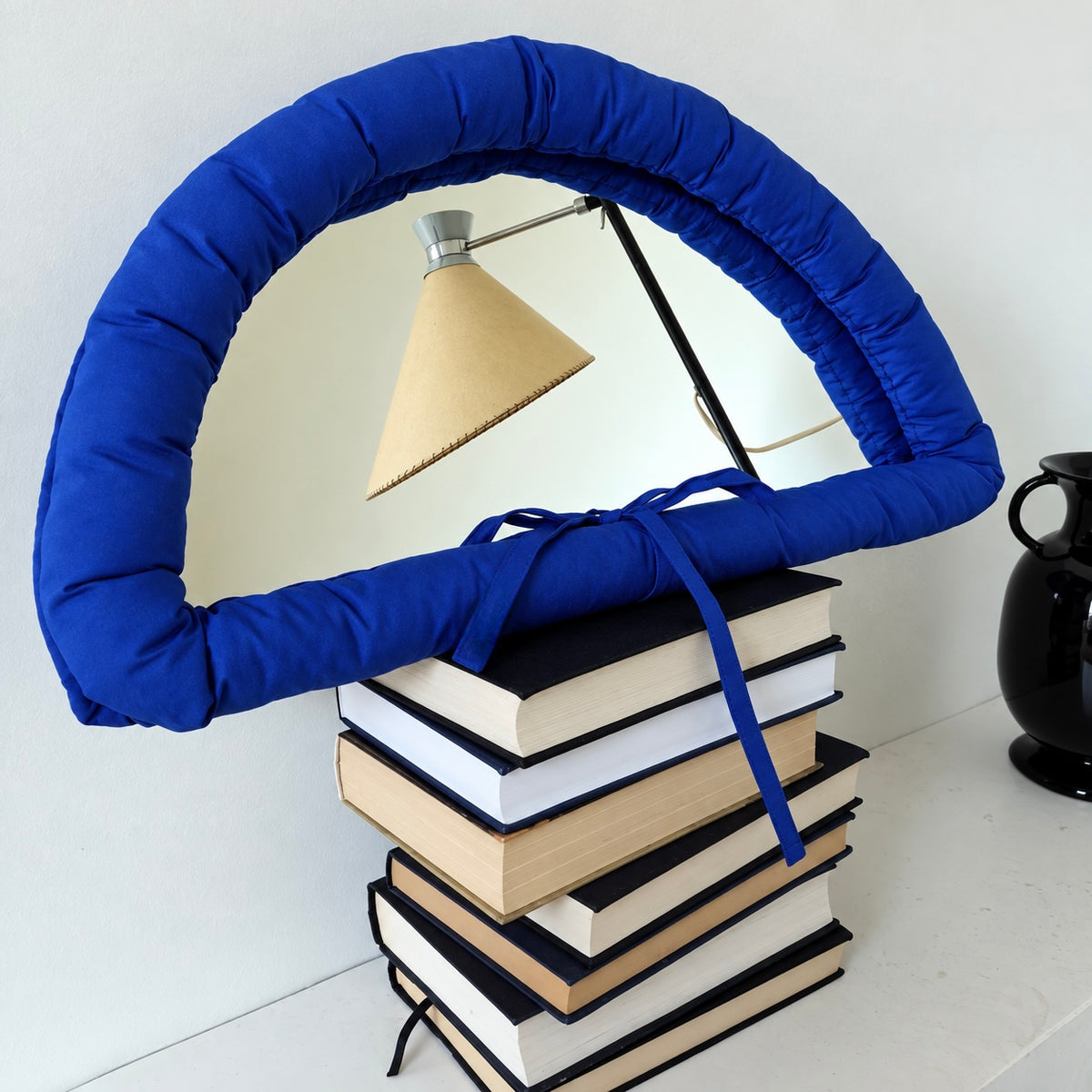 A blue padded arch-shaped lampshade or decorative lamp frame made of soft fabric, draped over a stack of books with a small table lamp visible behind it, against a plain light background.