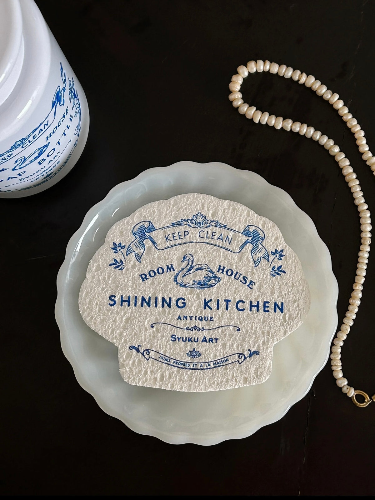 A scalloped dish sponge lies on a dark surface, showing blue swan artwork and lettering across the front.