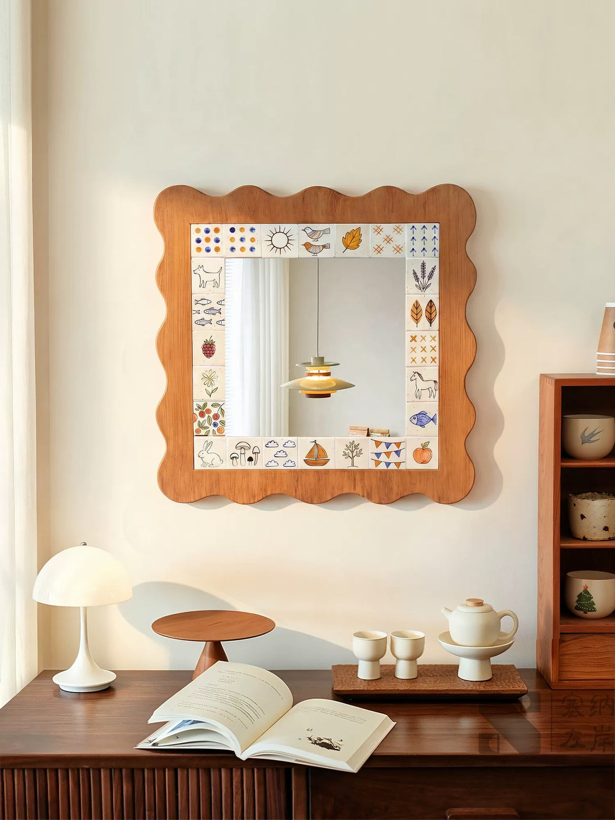 A scalloped mirror with a brown outer edge and illustrated border hangs above a cabinet with an open book and tea set.