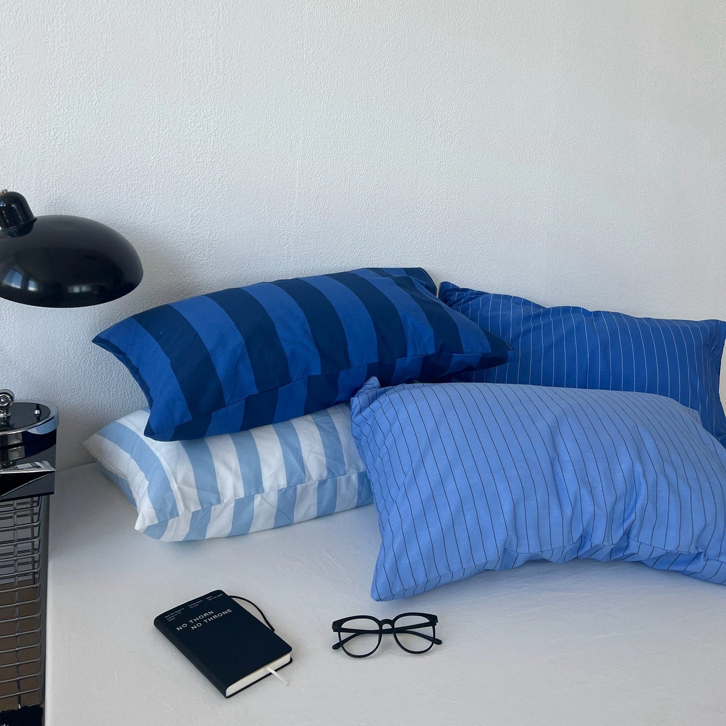 Four blue and navy striped and pinstriped pillows arranged on a bed, with a black desk lamp, notebook, and eyeglasses nearby against a white wall.