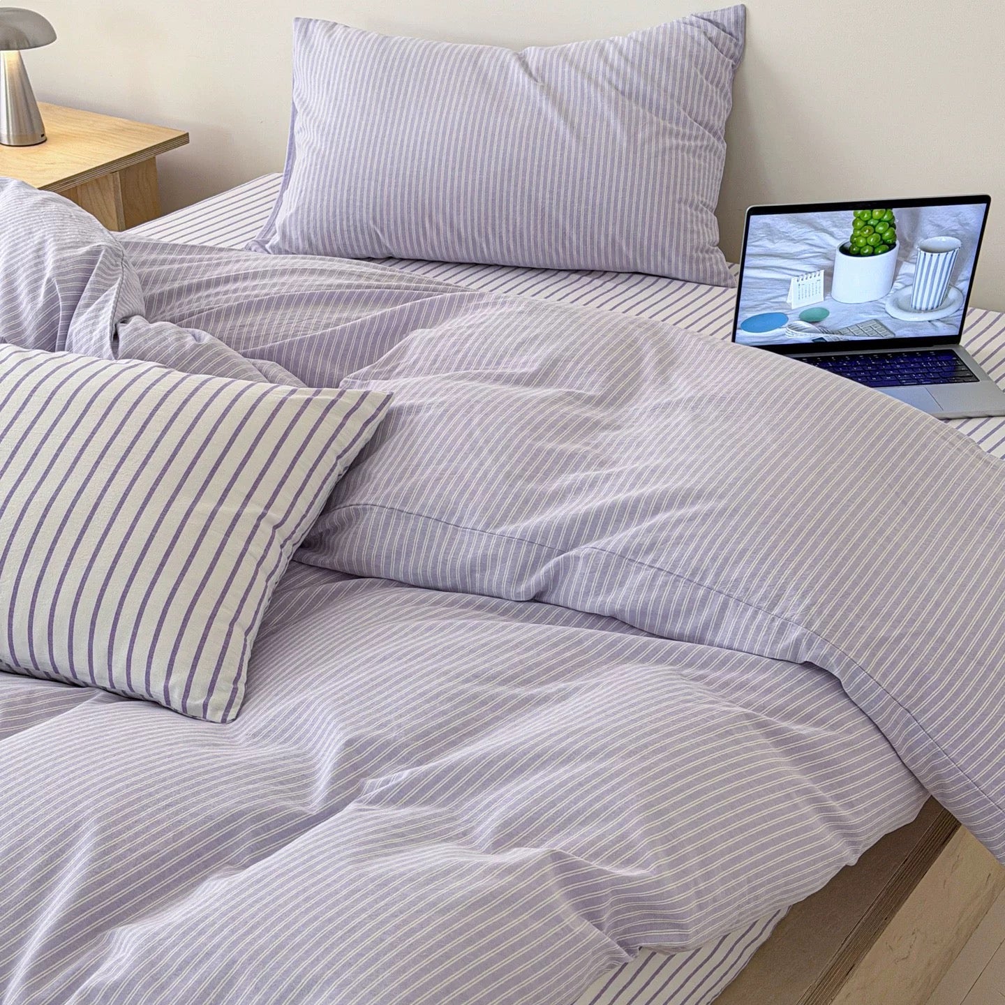 A bed dressed in light lavender and white vertically striped bedding, including a duvet cover and matching pillowcases, with a laptop open on the bedside table.