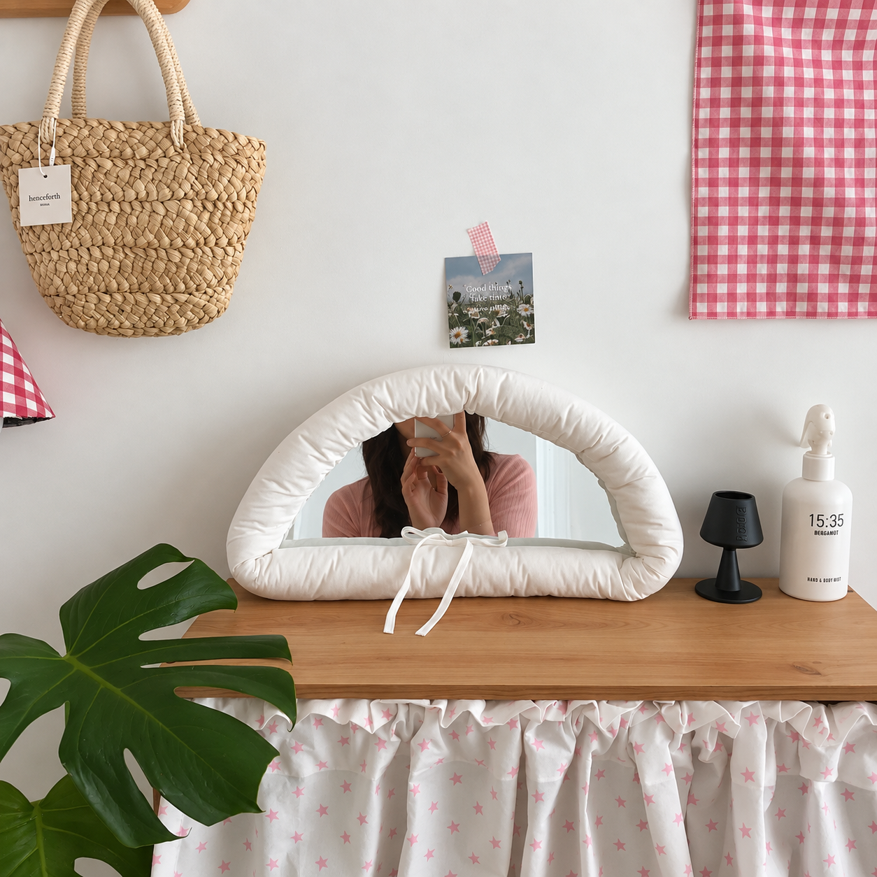 A white padded vanity mirror with a rounded arch frame and white tie details, styled on a wooden table in a room setting with a pink gingham curtain, a woven straw bag, and a green monstera leaf.