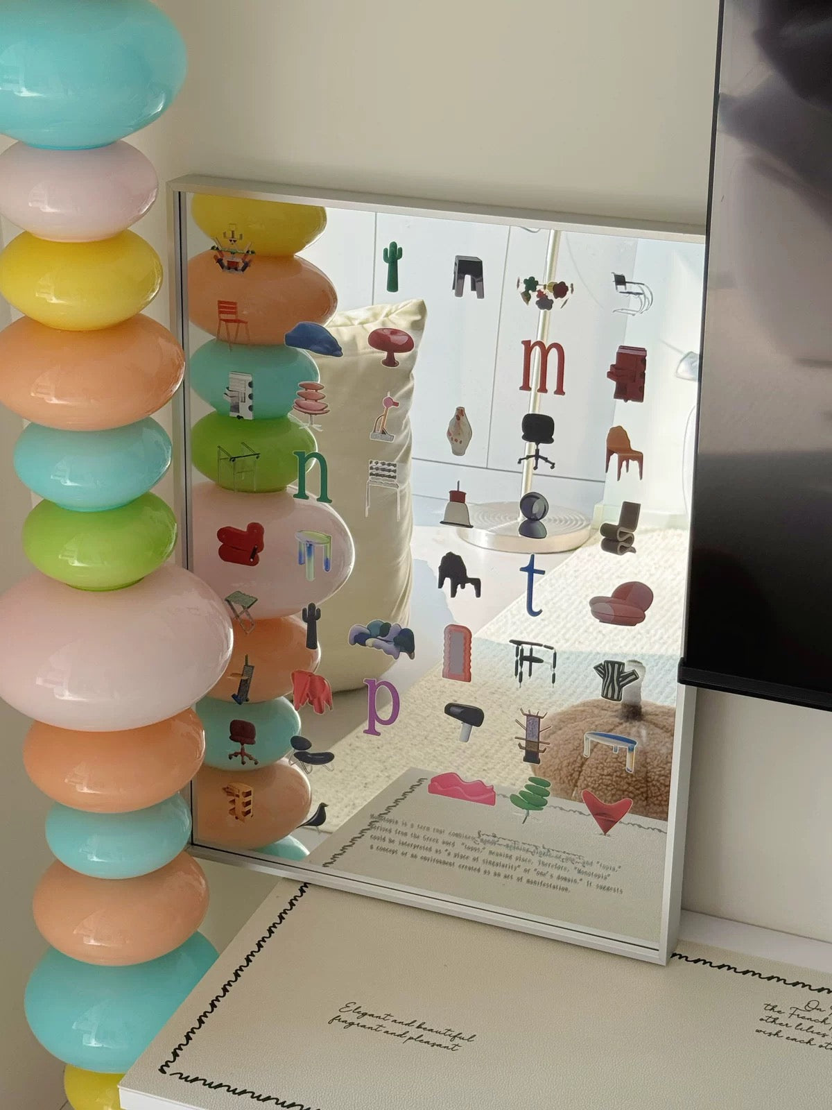 A square mirror with a white frame leaning against a wall, reflecting a room interior with small furniture icons and letters. A tall stack of pastel round objects in turquoise, pink, yellow, peach, and green stands to the left, and part of a black screen is visible on the right.