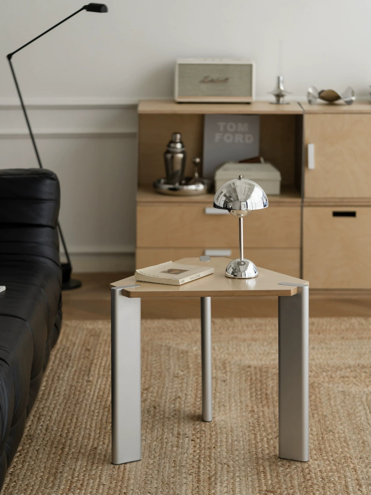 Silver triangle side table with a natural wood top and three broad metal legs holding books and a lamp.
