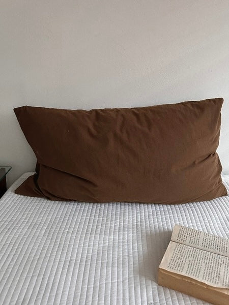 A single pillowcase in dark mocha brown rests upright on white bedding.