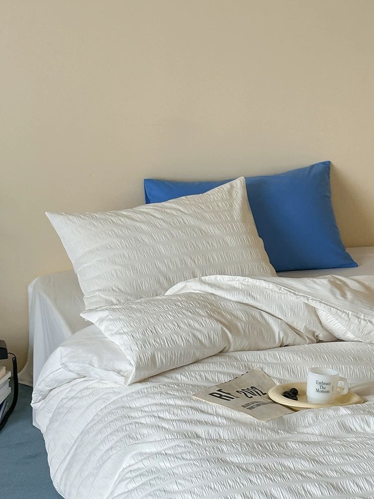 A bed styled with white textured bedding and two blue pillows against a plain beige wall, with a small tray and mug on the duvet.