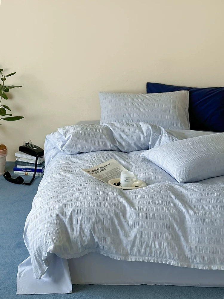A neatly made bed with light blue textured bedding, including a duvet cover and two pillows, styled in a bedroom setting with a dark blue pillow, a small plant, and a tray on the floor.