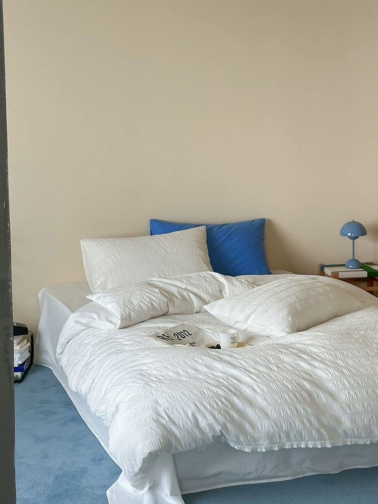 A bed dressed in white textured bedding with two white pillows and one blue accent pillow, set against a plain beige wall in a bedroom.