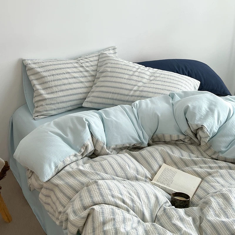 Bed with light blue bedding and striped pillows against a white wall.