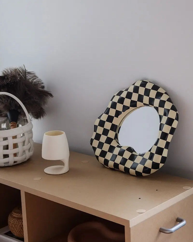 A scalloped-edge oval mirror with a black and cream checkerboard pattern, styled on a light wood cabinet next to a white ceramic vase and a basket with feathers.