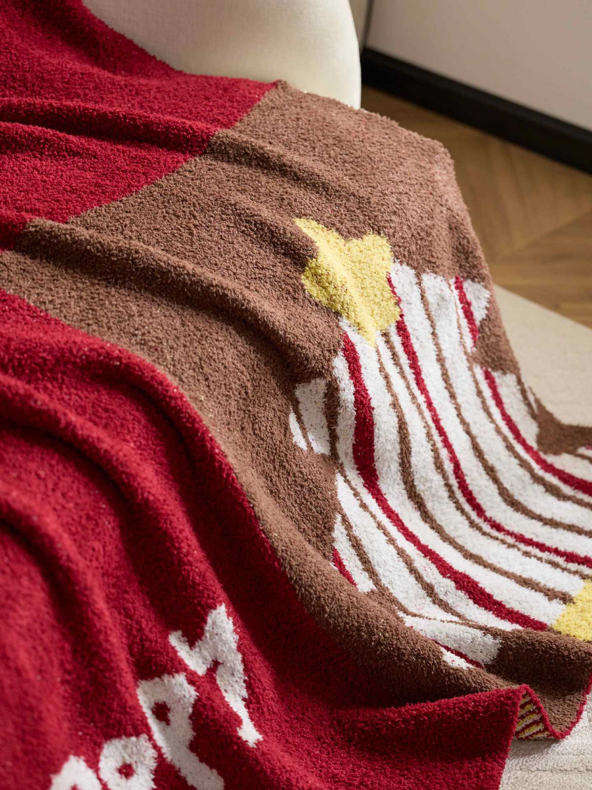 A close-up of a plush knit blanket draped over a white chair in a real indoor setting, showing a red and brown color-blocked design with white stripes and yellow star accents.