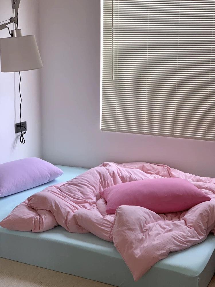 Bed with pink bedding and pillows in a room with a window and lamp.