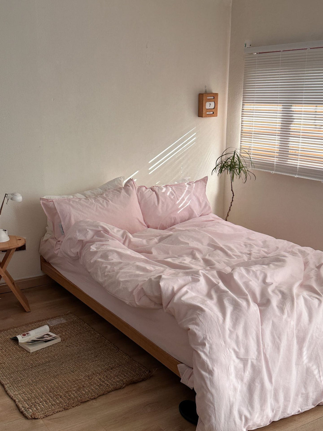A bed dressed in light pink bedding, including a duvet cover and two matching pillows, styled in a minimalist bedroom with a wooden side table, white lamp, and woven rug.