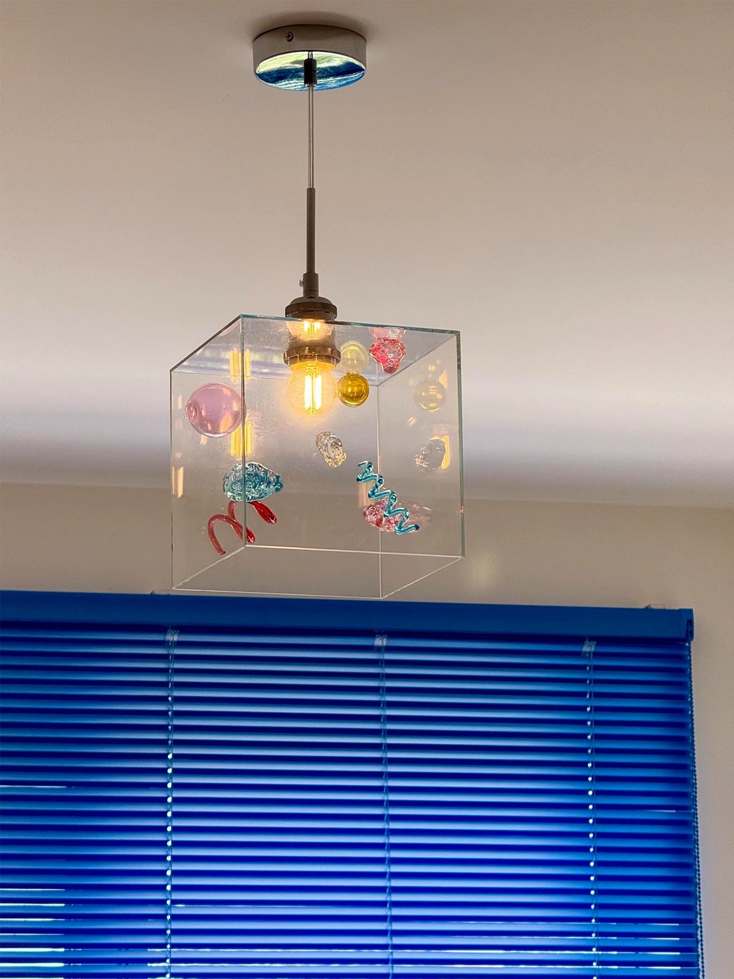 Modern pendant light with decorative elements in a clear cube, hanging above blue blinds.