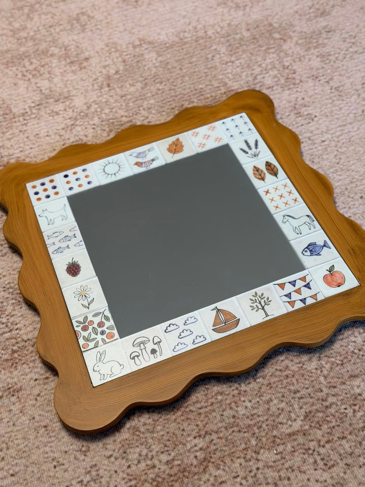 A square scalloped mirror with a brown frame lies flat on a pink surface, showing the full illustrated border.