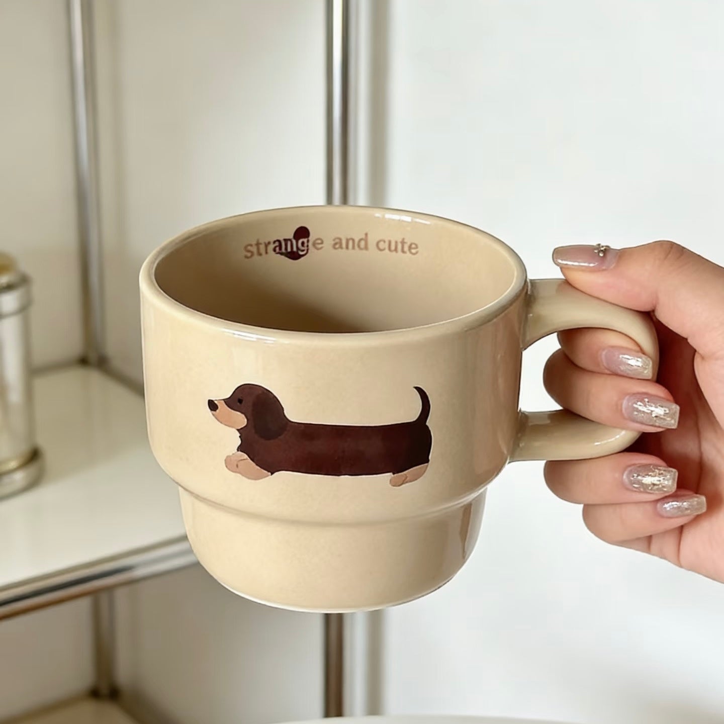 A hand with glittery nail polish holds a beige ceramic mug featuring a brown dachshund illustration and the text 'strange and cute' inside the mug.