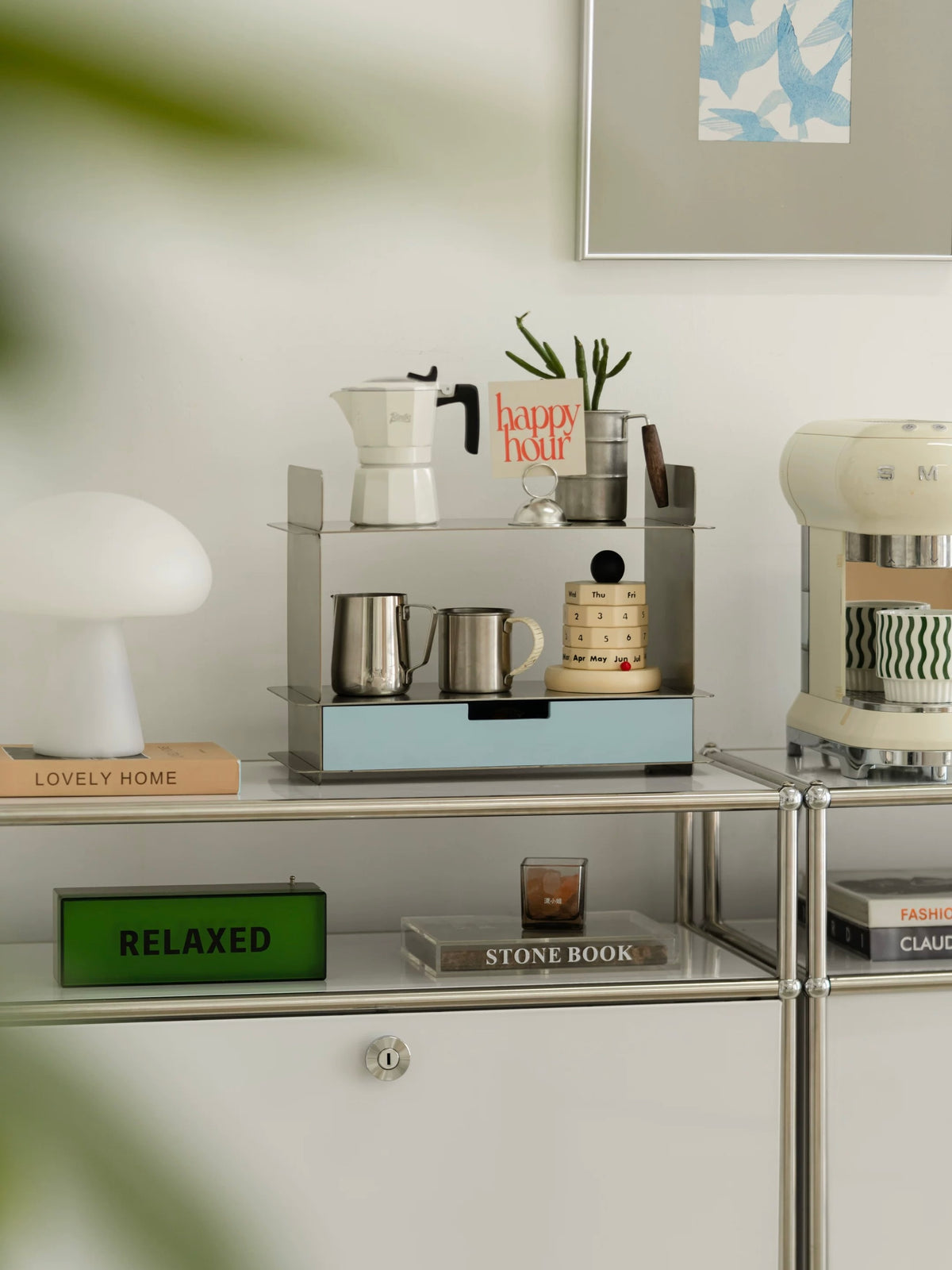 Stainless steel shelf with a drawer on a console beside a green plant and framed art