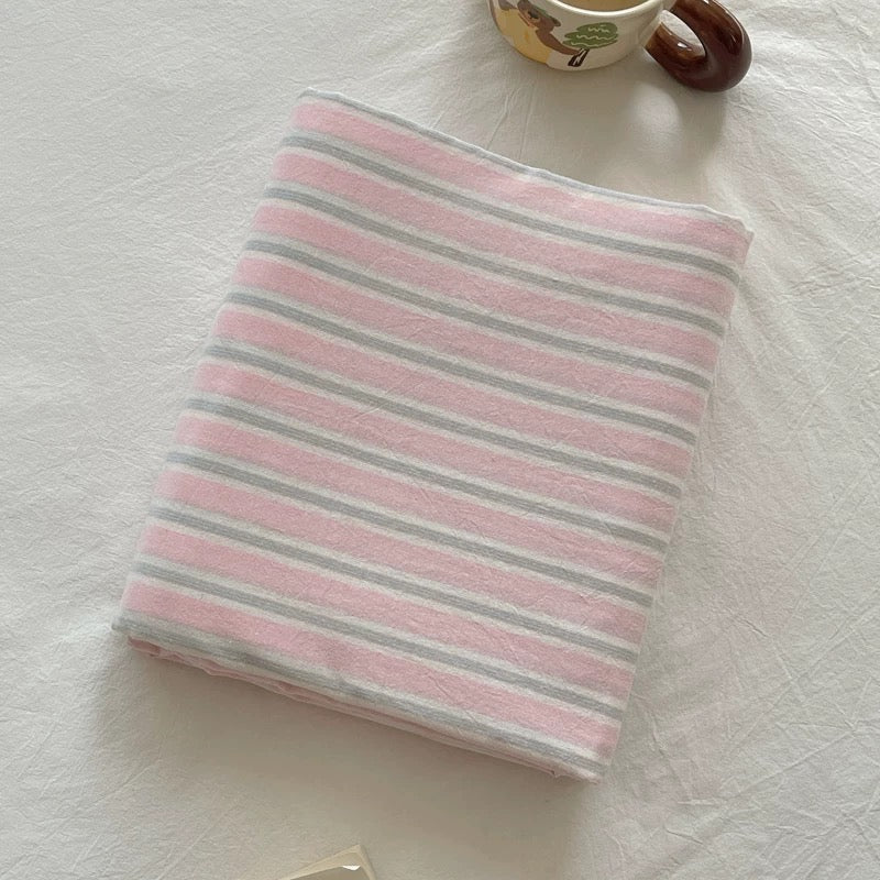 A stack of five folded striped textiles, likely towels, blankets, or sheets, in soft pastel colors including pink, blue, yellow, and green on a white base. A small glass jar with a white label sits on top.