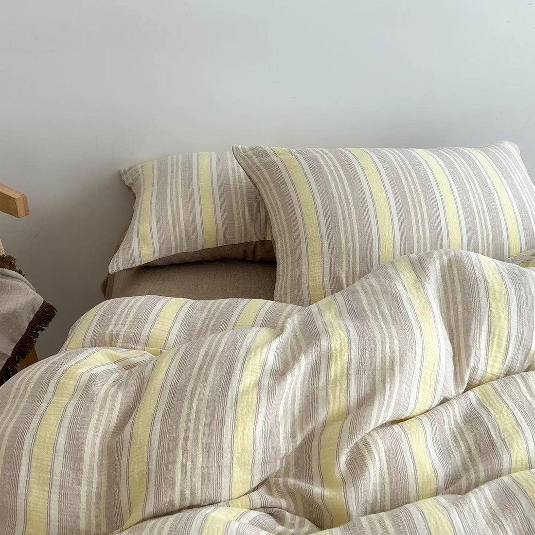A cozy bed styled with a beige and white striped bedding set, including two striped pillows, a matching duvet cover, and solid beige pillows, set against a plain white wall.