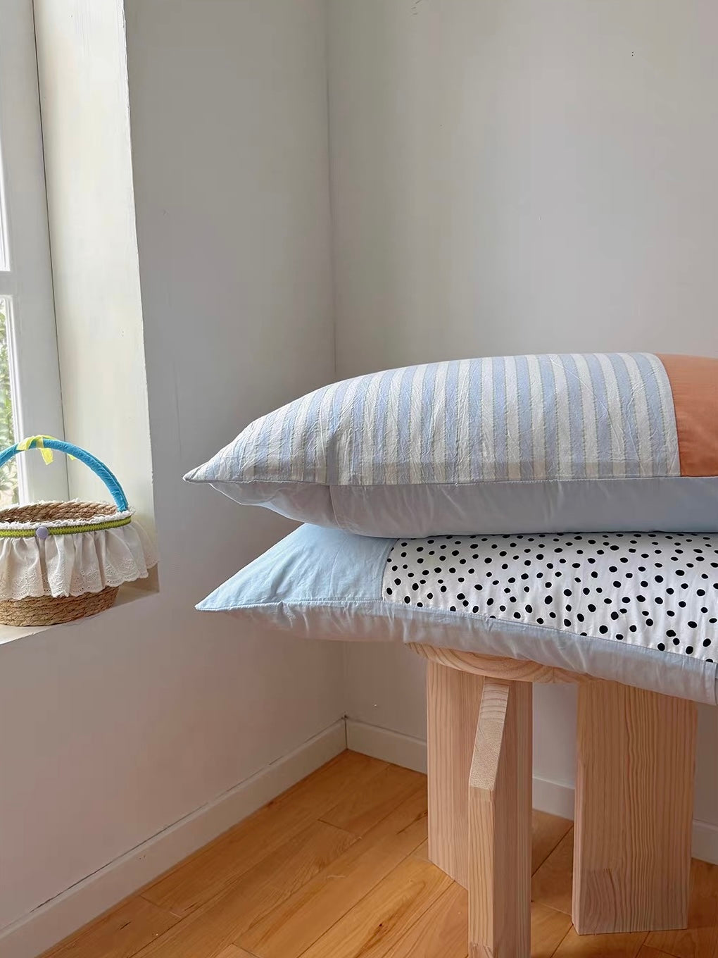 A striped pillowcase with a peach panel rests above a blue and dotted pillowcase on a wooden stool.
