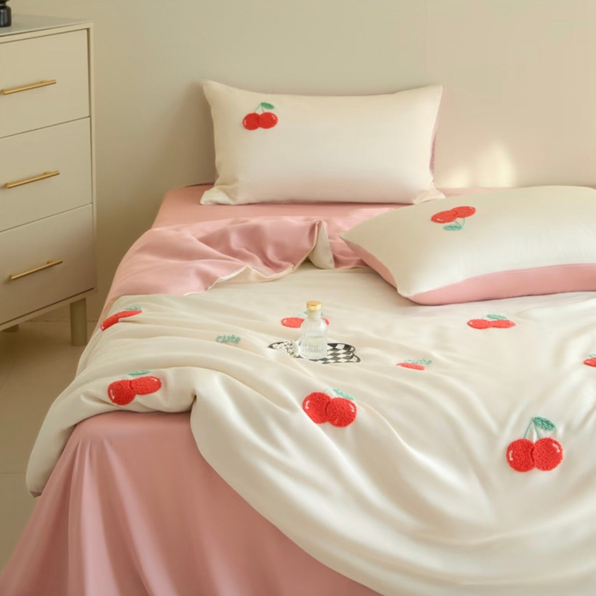 A bed dressed in a soft pink and off-white bedding set with embroidered red cherries, shown in a bedroom setting next to a white dresser.