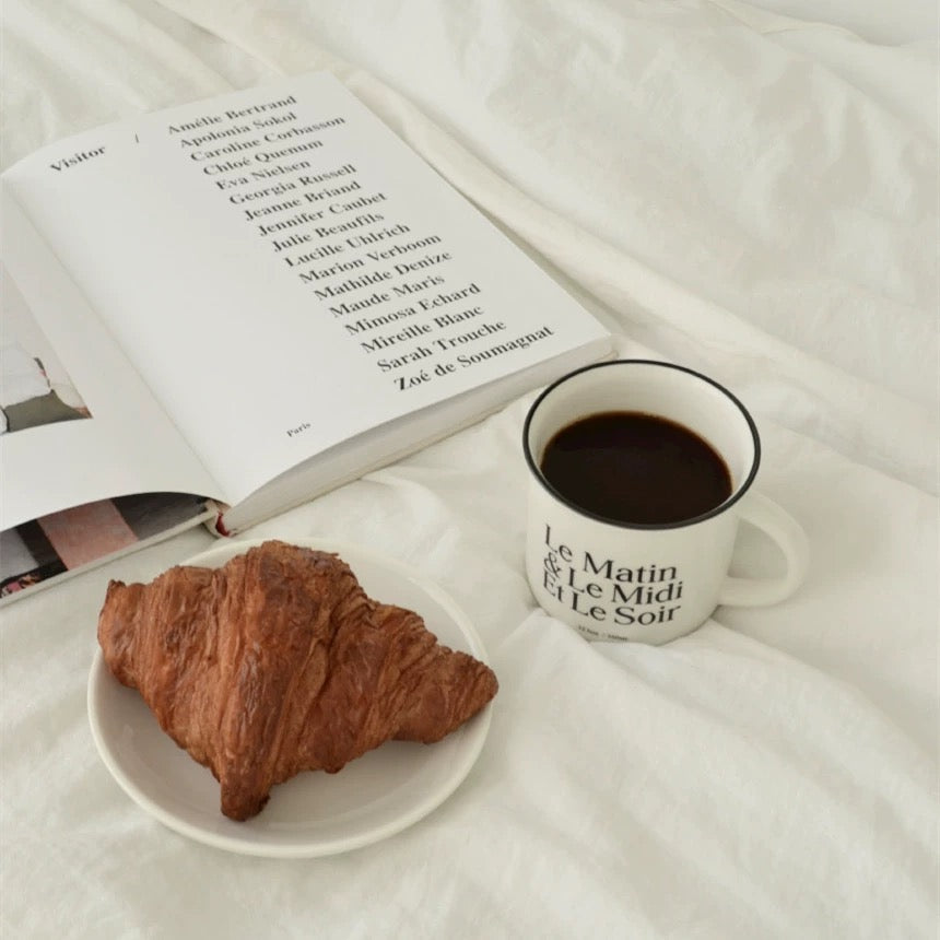 A white ceramic mug filled with black coffee sits on a white plate next to a golden-brown croissant on a white bedspread, with an open book in the background.