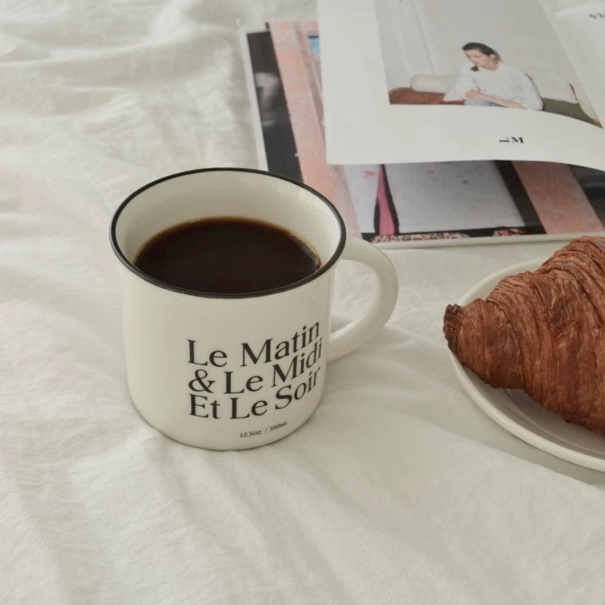 A white ceramic mug filled with black coffee sits on a white fabric surface, printed with the French text 'Le Matin & Le Midi Et Le Soir'. A croissant on a plate is partially visible at the right, with a magazine open behind the mug.